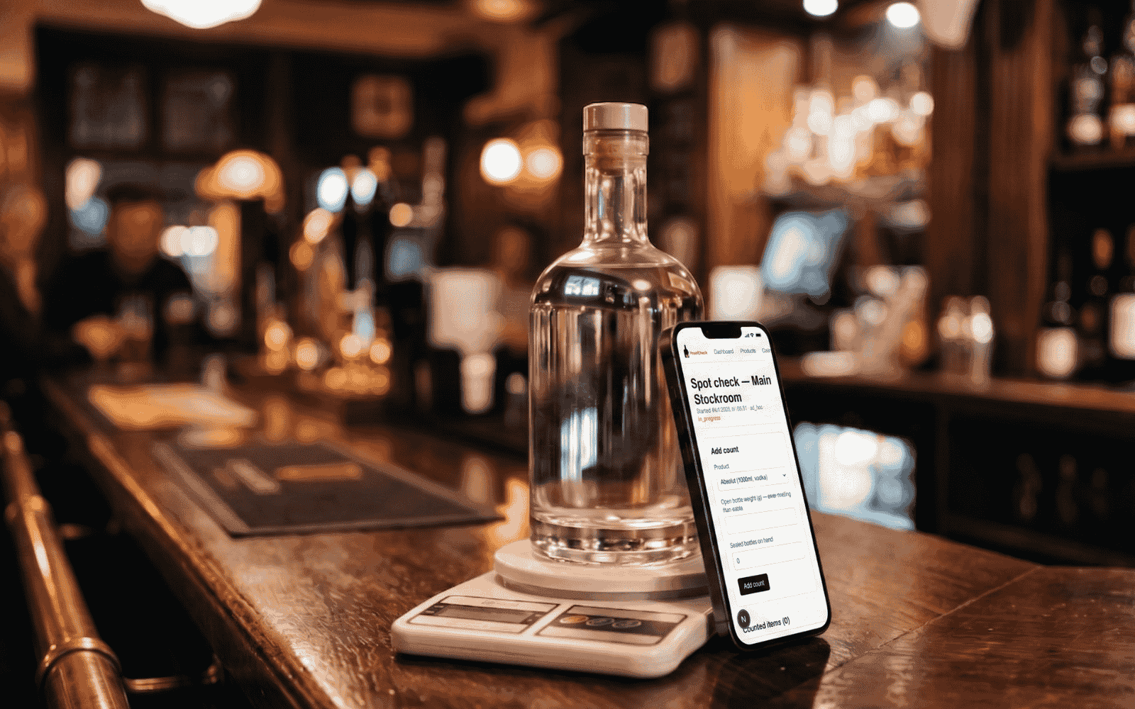 A spirits bottle on a digital kitchen scale with a phone leaning against it on a polished bar top.